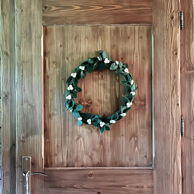 Couronne de Noël artisanale gui et houx New Heidi suspendue sur une porte en bois rustique.