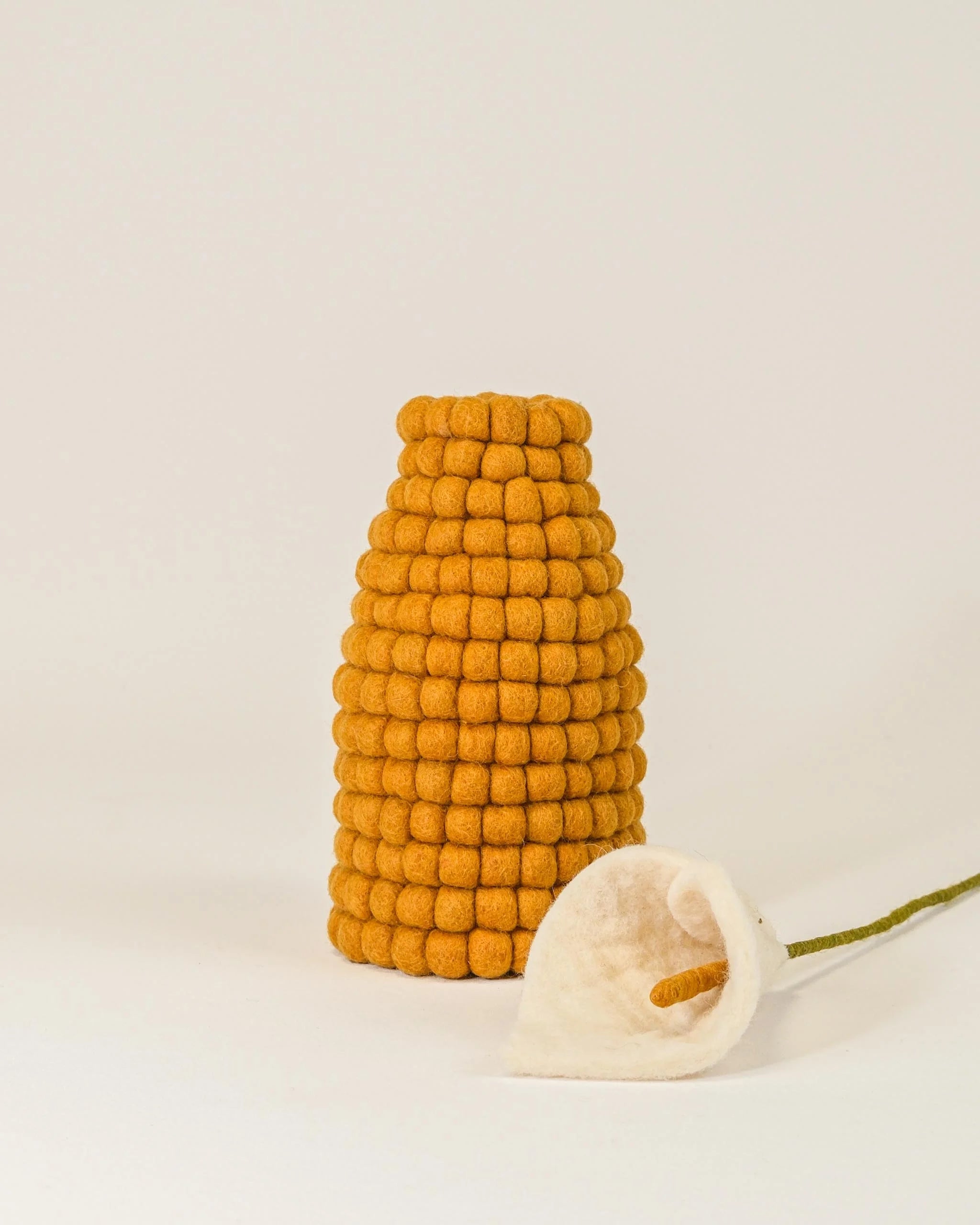 Photo d'un vase en feutrine moutarde sur un fond blanc avec une fleur d'arum à coté.