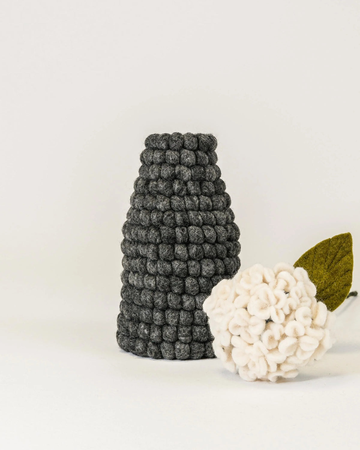 Photo d'un vase en feutrine gris sur un fond blanc avec une hortensia à coté.
