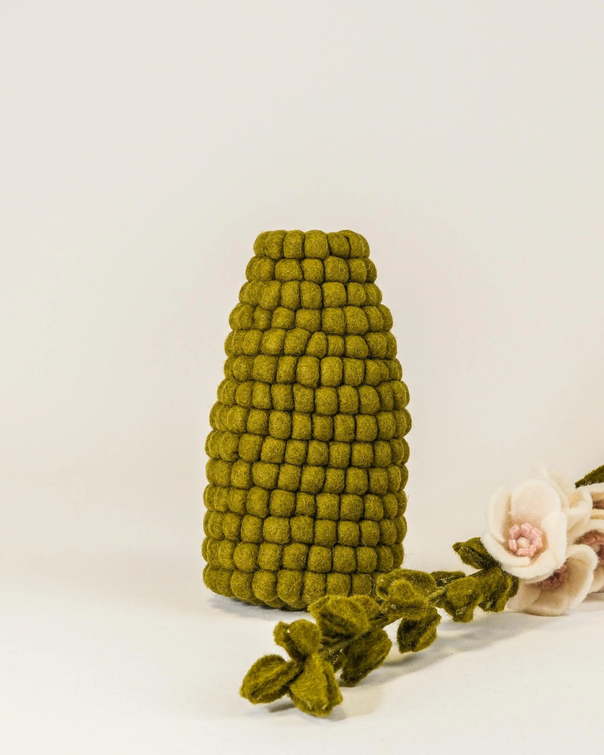 Vase en feutrine kaki sur un fond blanc avec une rose tremiere blanche a coté.