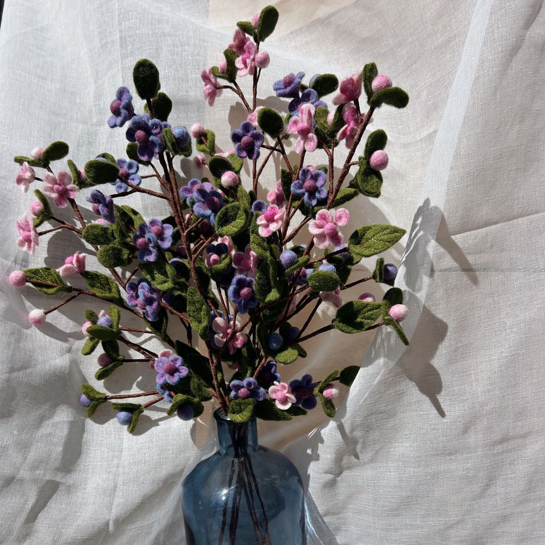 Composition florale printanière avec des branches de fleurs en laine feutrée roses, blanches et mauves dans un flacon bleu.