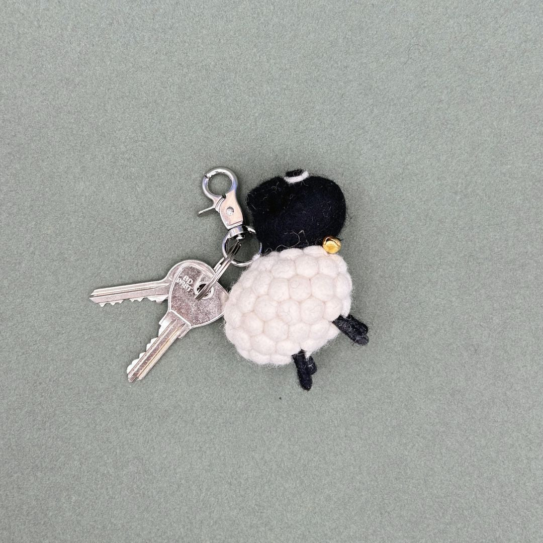 Porte-clé en feutrine en forme de mouton à tête noire avec son petit grelot doré et son trousseau de clés sur fond gris.