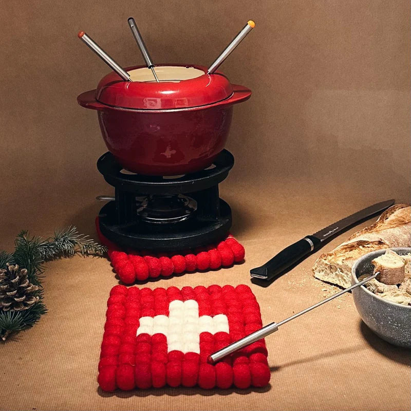 Caquelon à fondue rouge posé sur le dessous de plat drapeau suisse New Heidi, décor montagne.