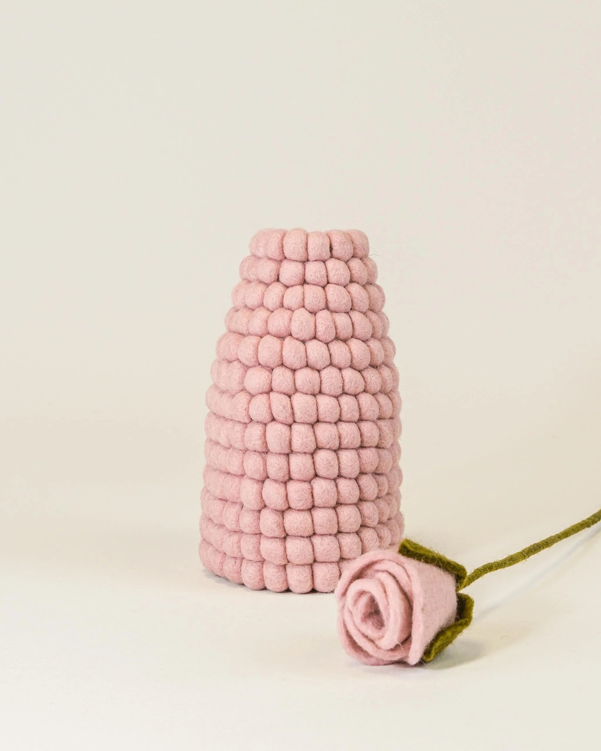 Photo d'un vase en feutrine rose sur un fond blanc avec une rose à coté. 