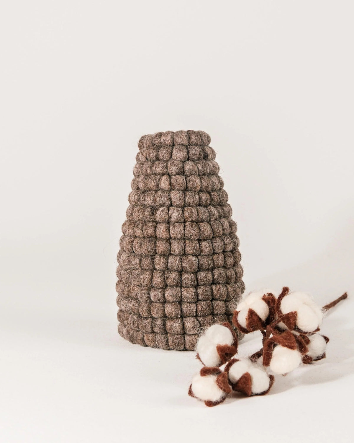 Photo d'un vase en feutrine marron sur un fond blanc avec une fleur de coton à coté. 