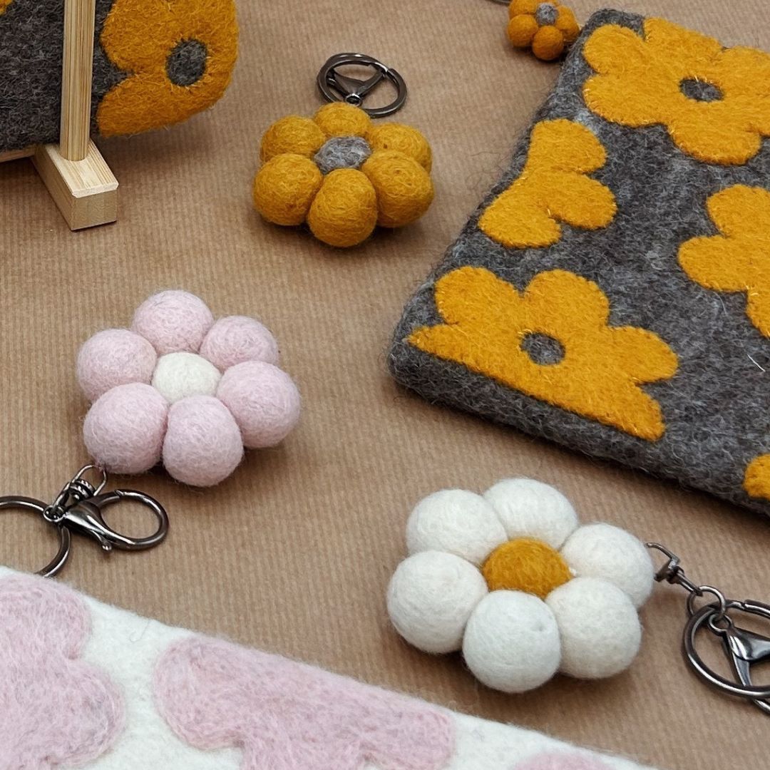 Collection de porte-clés fleurs New Heidi en laine feutrée moutarde, rose et blanche posés à côté d'une pochette en feutrine.