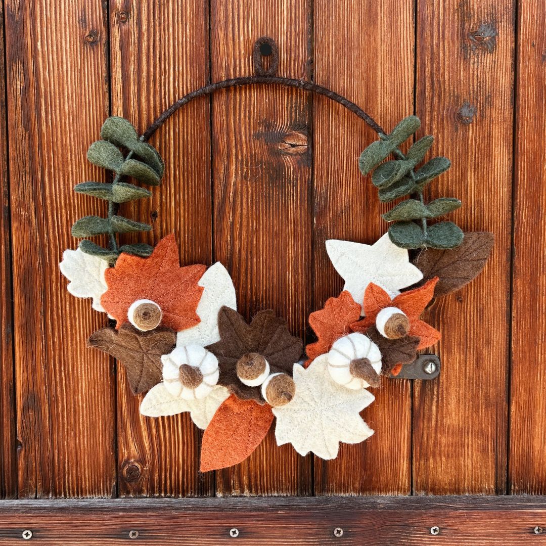 Couronne d'automne de 35cm en feutrine New Heidi composée de petites feuilles ocre, citrouilles blanches et glands en laine, vue de trois-quarts sur un volet en bois.