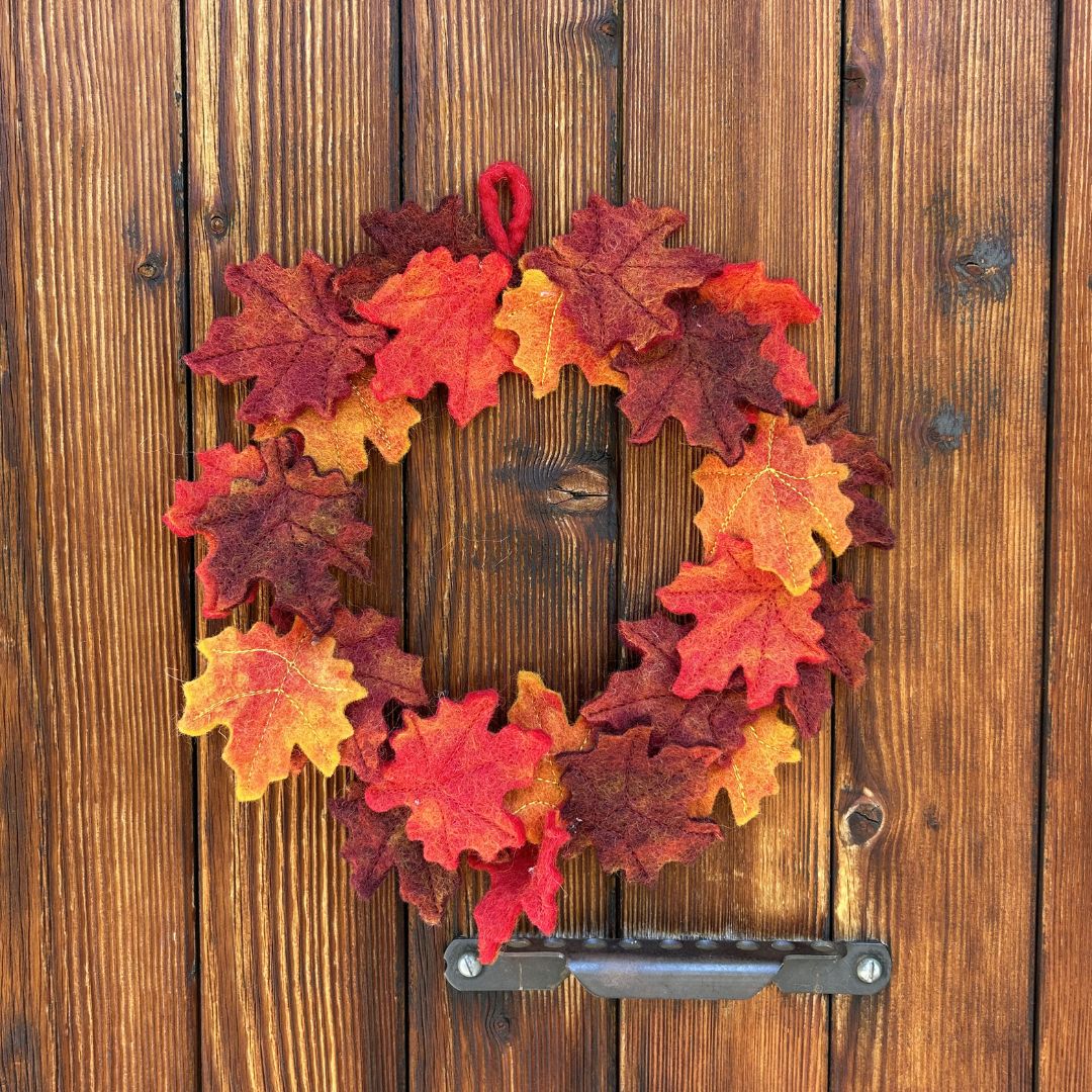 Couronne d'automne de 20cm en feutrine New Heidi composée de feuilles d'érable artisanales ocre, bordeaux et terracotta, suspendue de face sur une porte en bois.