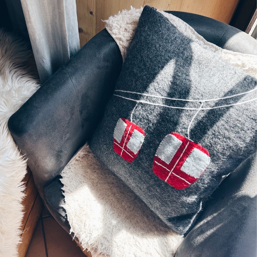 Coussin carré déhoussable en feutrine grise New Heidi avec un motif de télécabine rouge et blanche en laine feutrée.