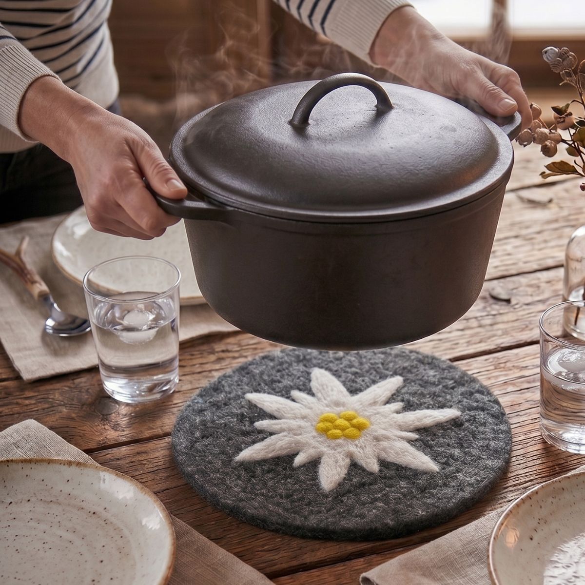 Personne posant une cocotte en fonte chaude sur un dessous de plat edelweiss en laine feutrée sur une table en bois.