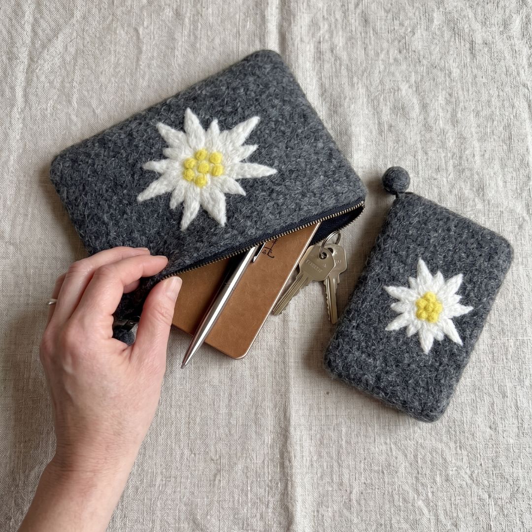 Grande pochette edelweiss ouverte contenant un carnet et un stylo à côté d'un petit étui de téléphone en laine bouillie.
