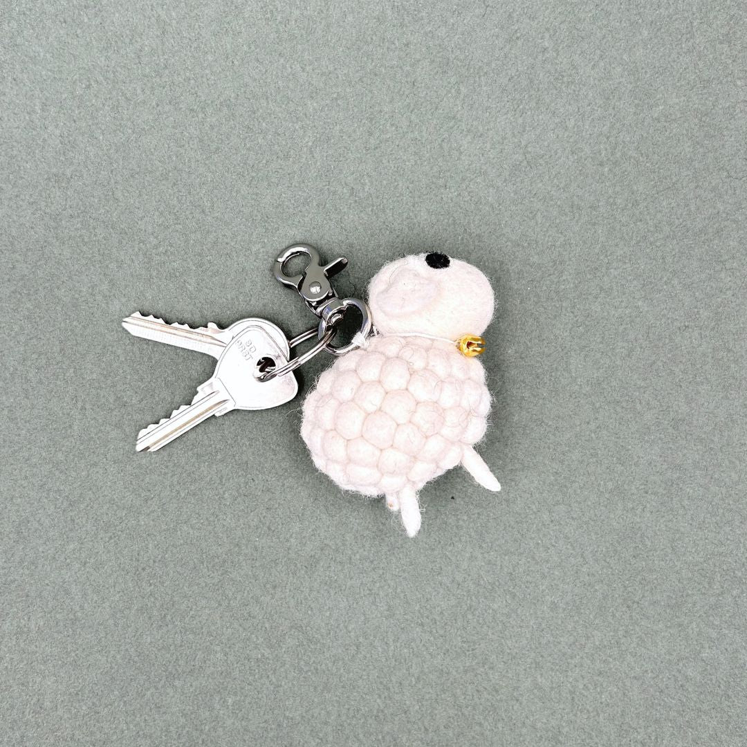 Porte-clé en feutrine en forme de petit mouton blanc avec grelot doré et son trousseau de clés sur fond gris.