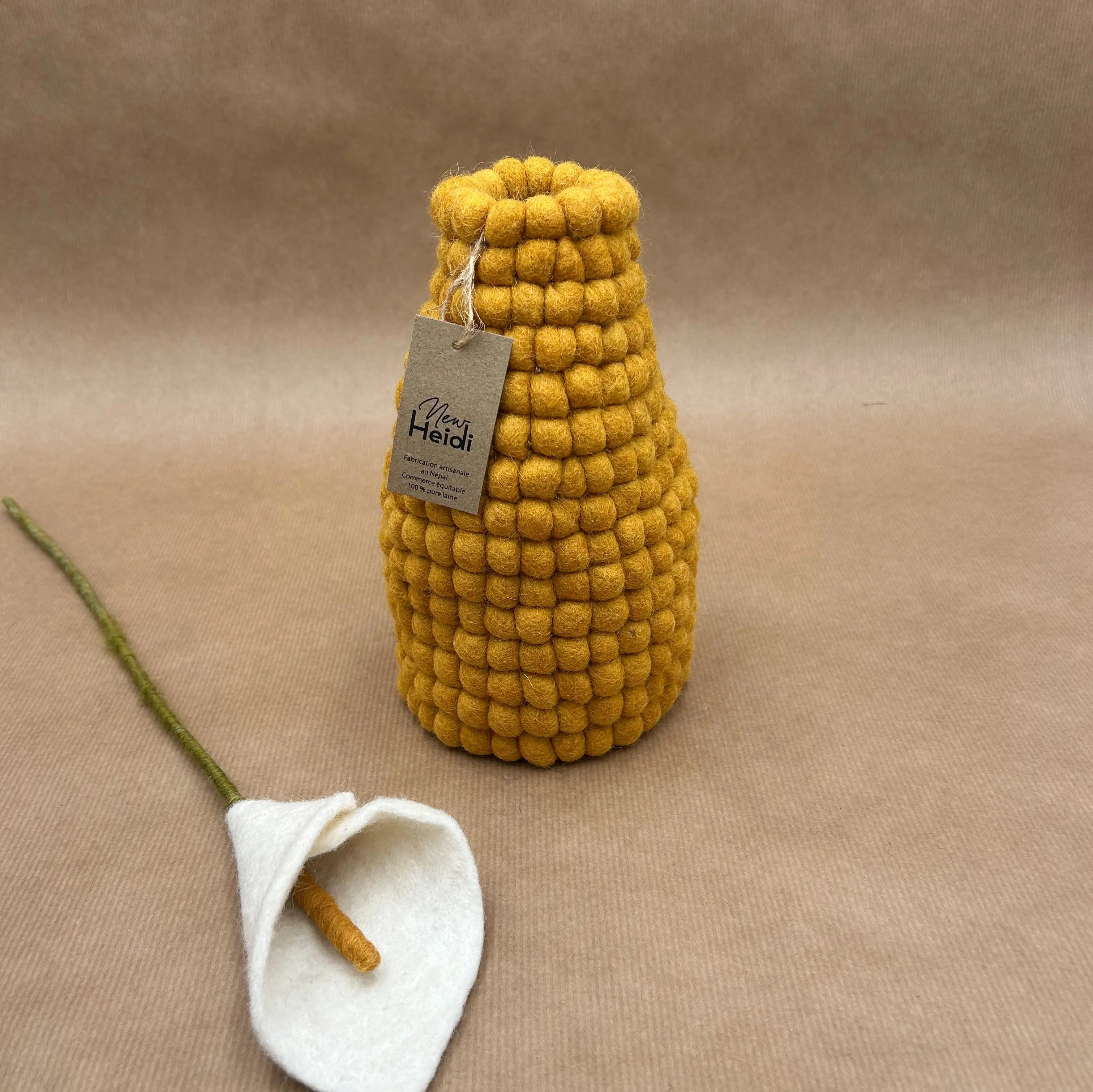 Vase en laine feutrée moutarde New Heidi avec son étiquette artisanale, posé à côté d'une fleur d'arum blanche.